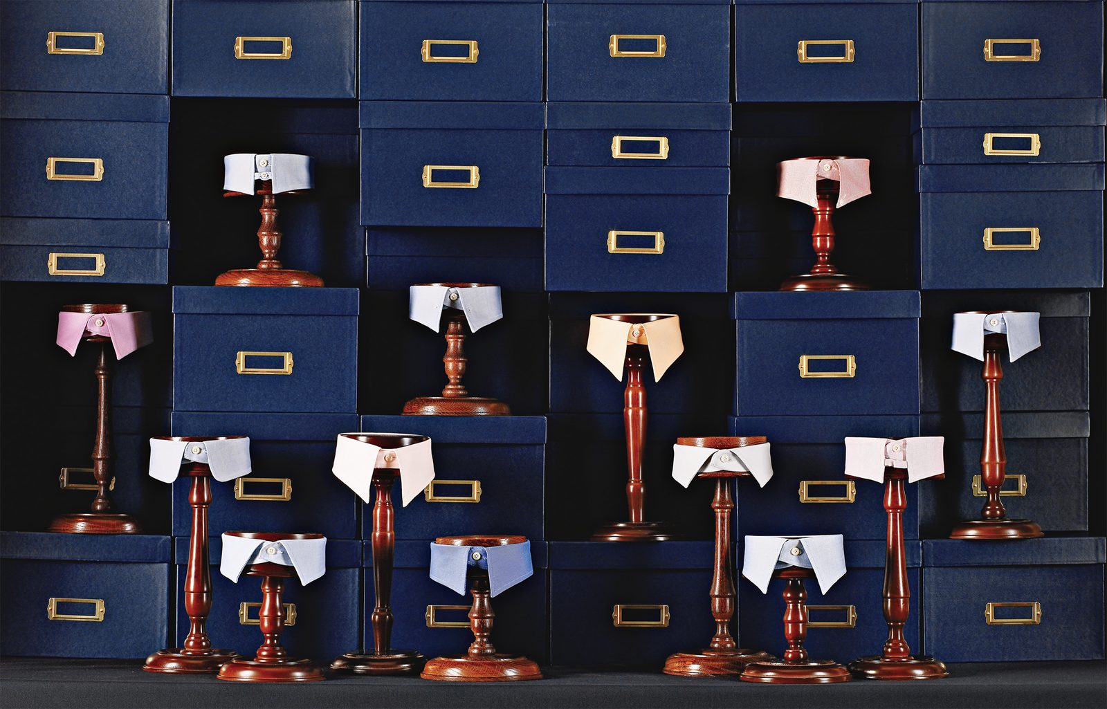Fourteen shirt collars on turned wooden stands against a wall of navy shirt boxes.