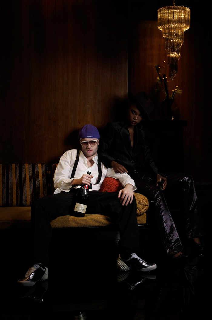 A man in a purple cap on a banquette holding champagne, a woman in a fedora behind him.
